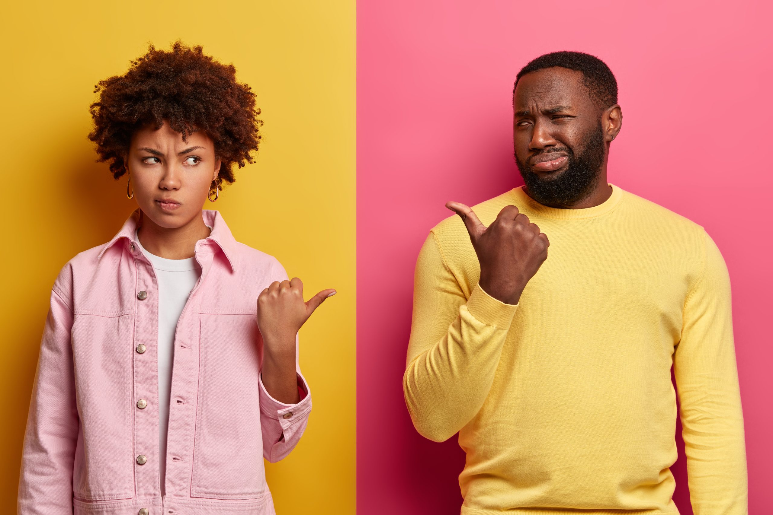 Photo of discontent pessimistic ethnic curly girlfriend and boyfriend point thumbs at each other, smirk faces unhappily, blame in doing something wrong, stand against yellow and pink studio wall